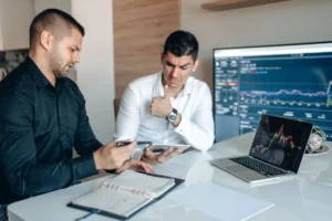 Two finance professionals reviewing financial data on a tablet with market charts displayed on laptops.